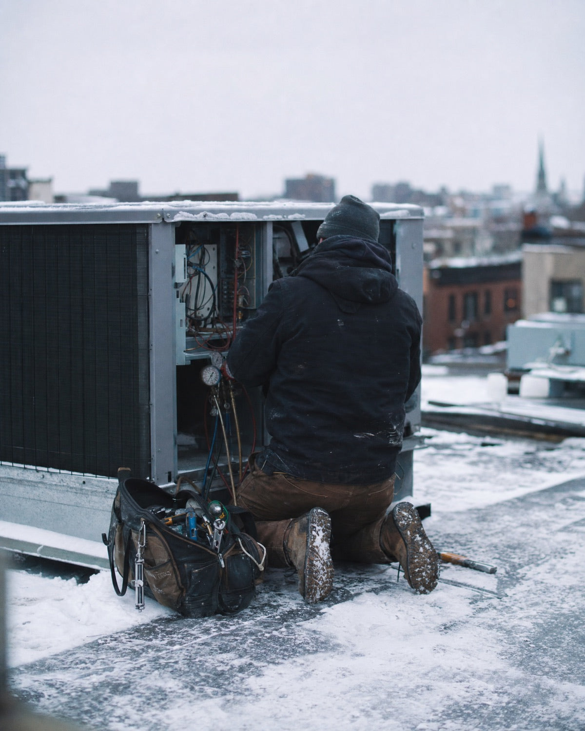 HVAC technician kneeling at a rooftop unit in the snow, tool bag open beside them