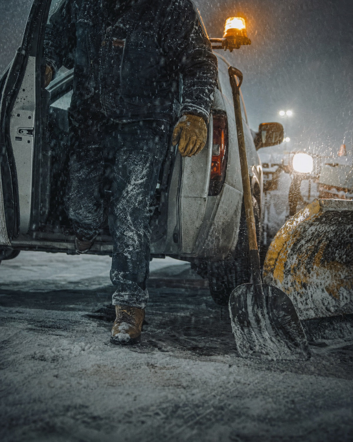 Plow operator stepping out of a truck into heavy snow at night, shovel leaning on the truck